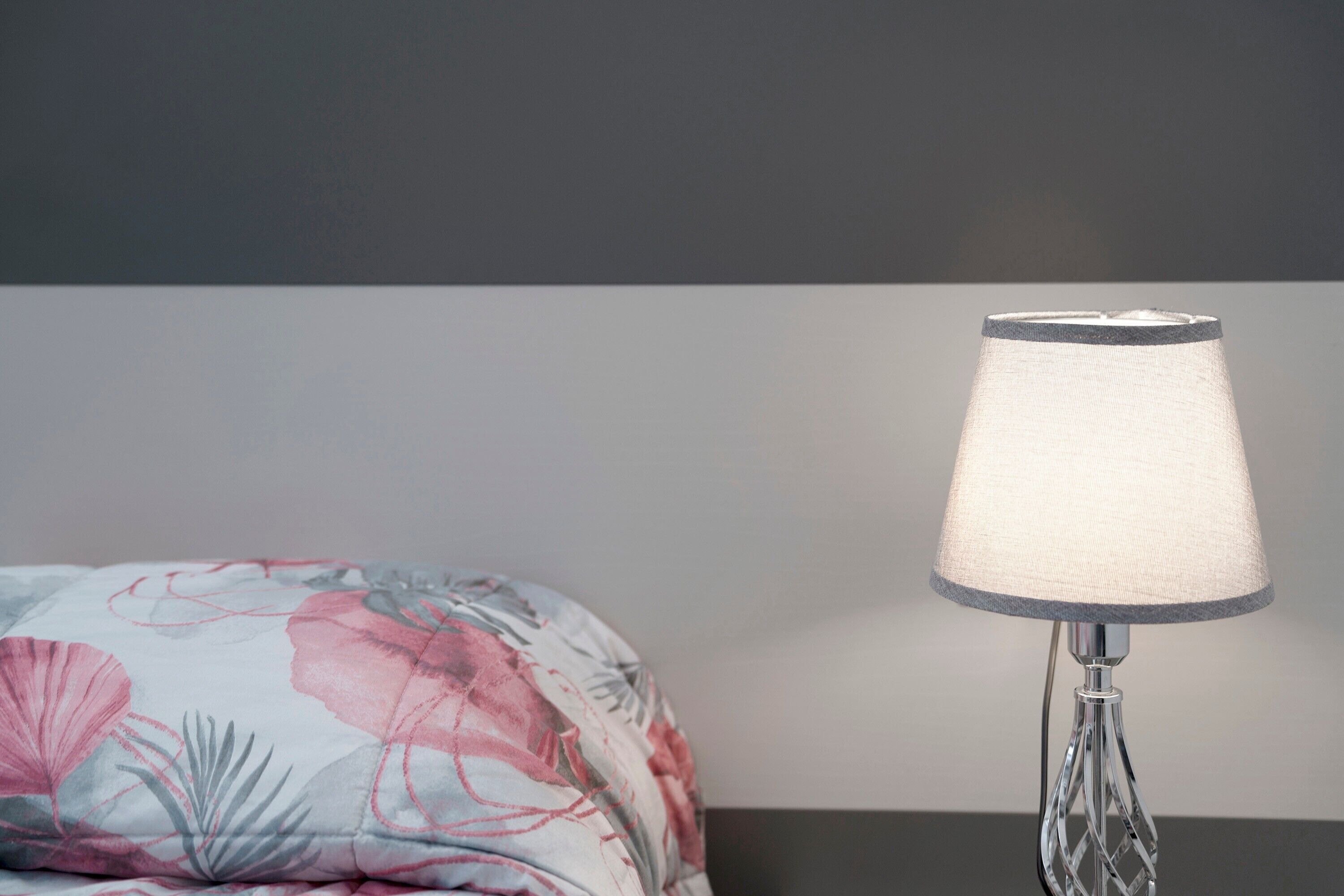 Un lit recouvert d’une couvre-lit à motifs floraux, une lampe moderne avec un abat-jour blanc et un mur sombre en arrière-plan.