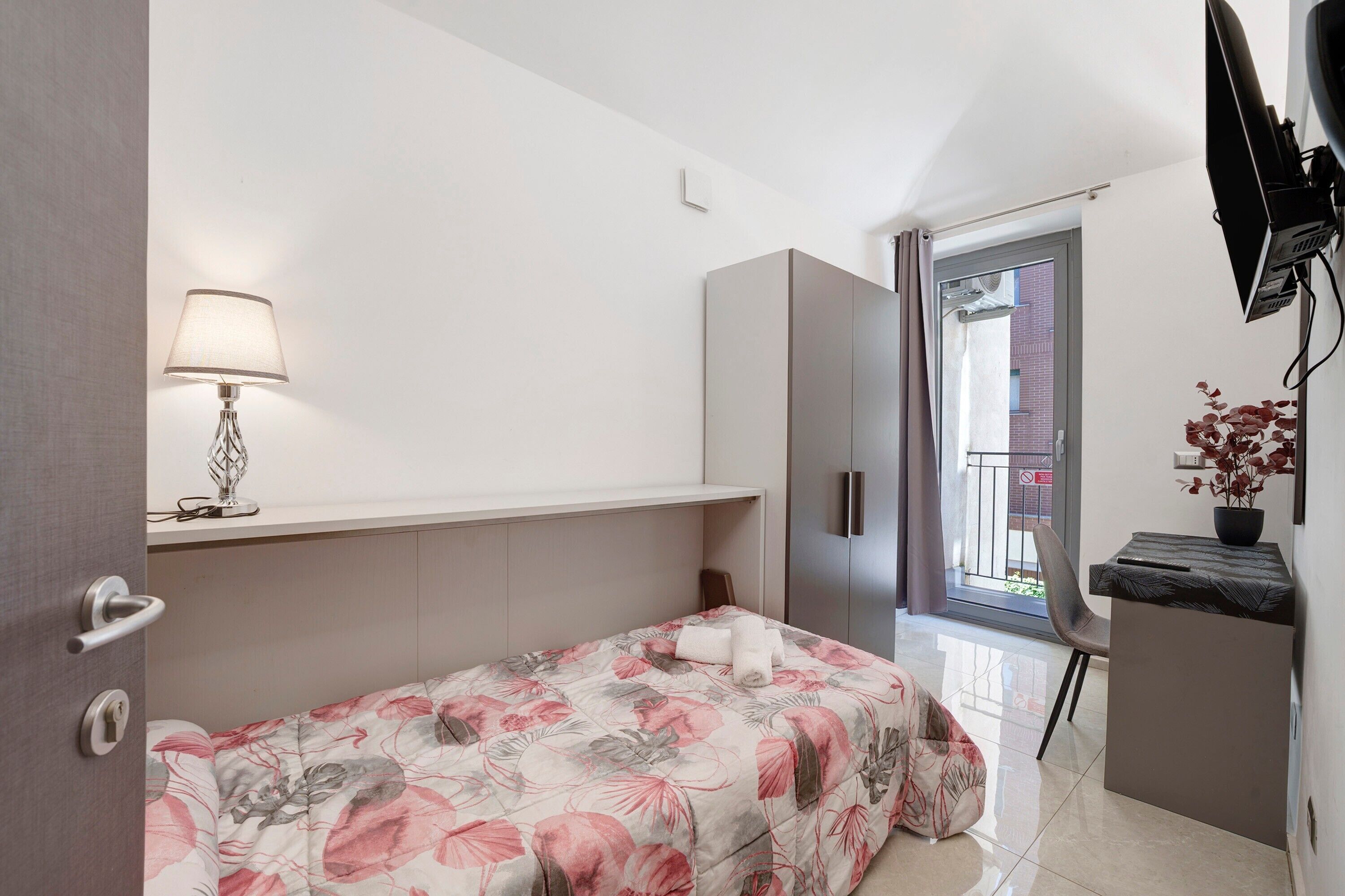 A bedroom with a bed, a desk, a lamp, and a TV.