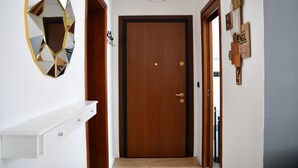 Um corredor com uma porta de madeira, um console branco, um espelho geométrico e um lustre.