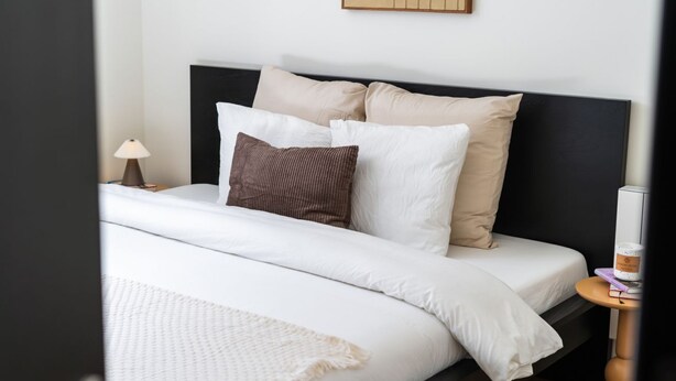 A neatly made bed with white and beige bedding, a brown pillow, and a small bedside table with a lamp.