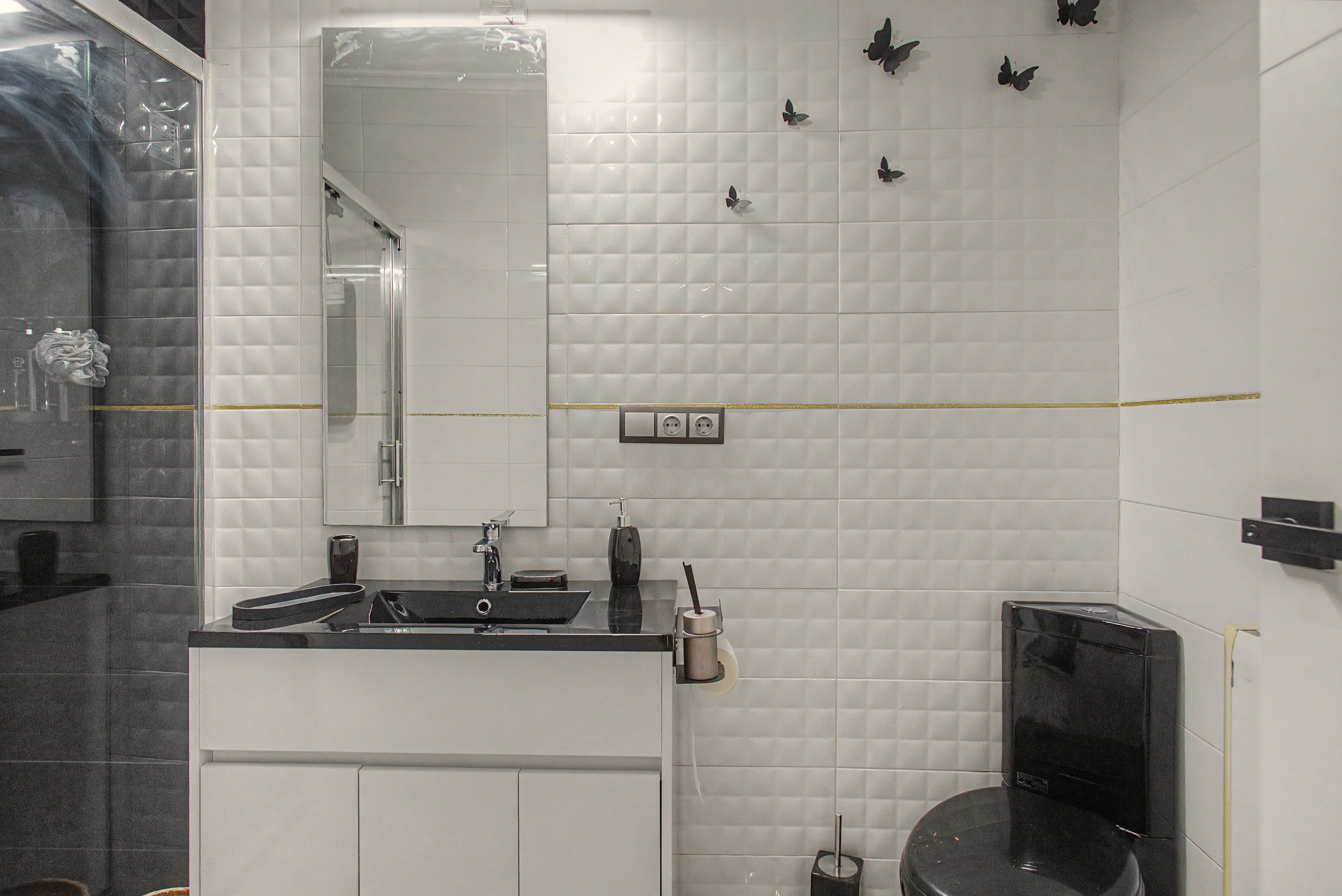 Un baño con pared de azulejos blancos, un espejo, un lavabo con encimera negra, un inodoro y una zona de ducha.
