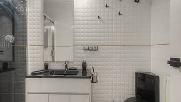 Un baño con pared de azulejos blancos, un espejo, un lavabo con encimera negra, un inodoro y una zona de ducha.