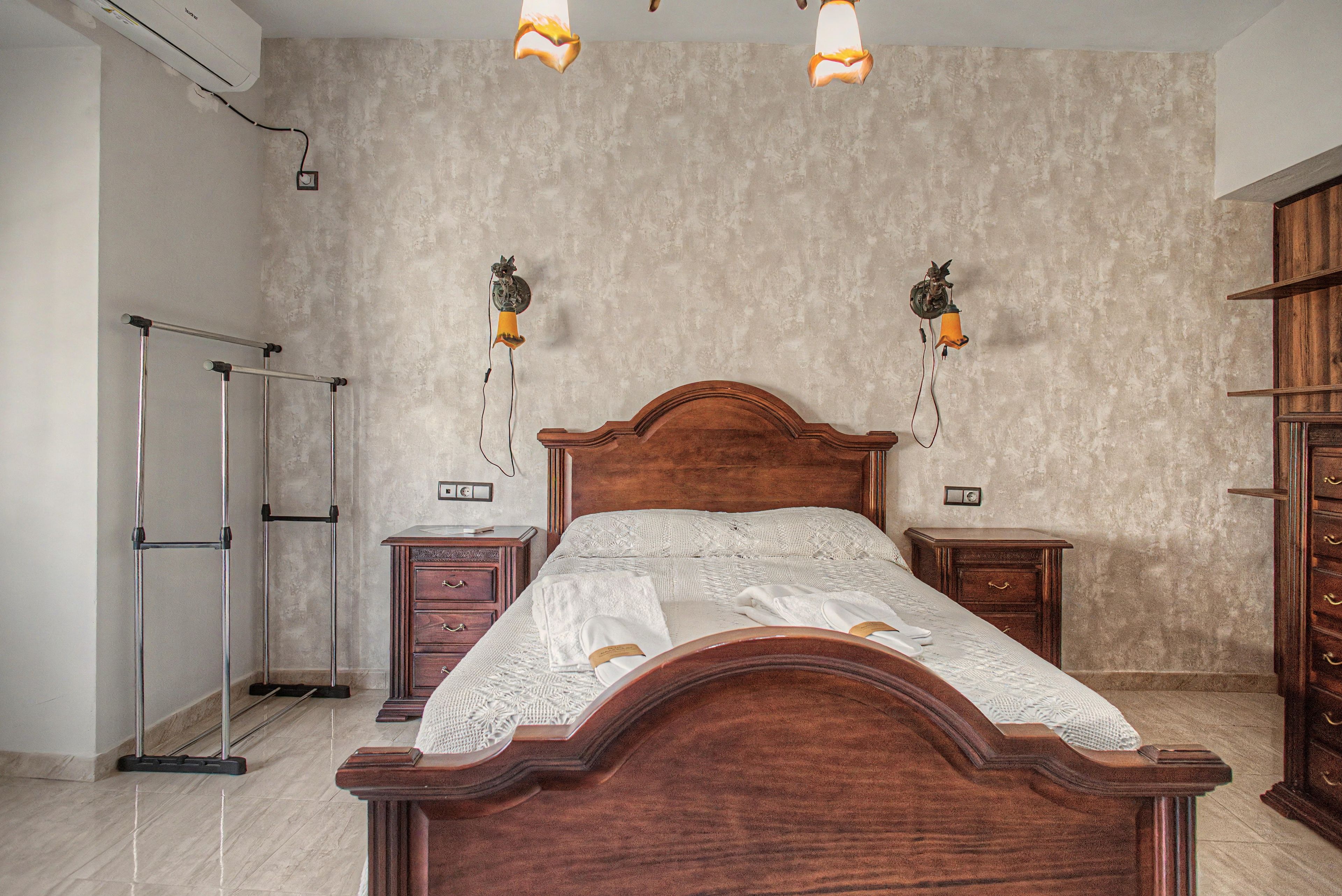 Une chambre à coucher avec un lit en bois, deux tables de chevet, une commode, une étagère fixée au mur et un ventilateur de plafond.