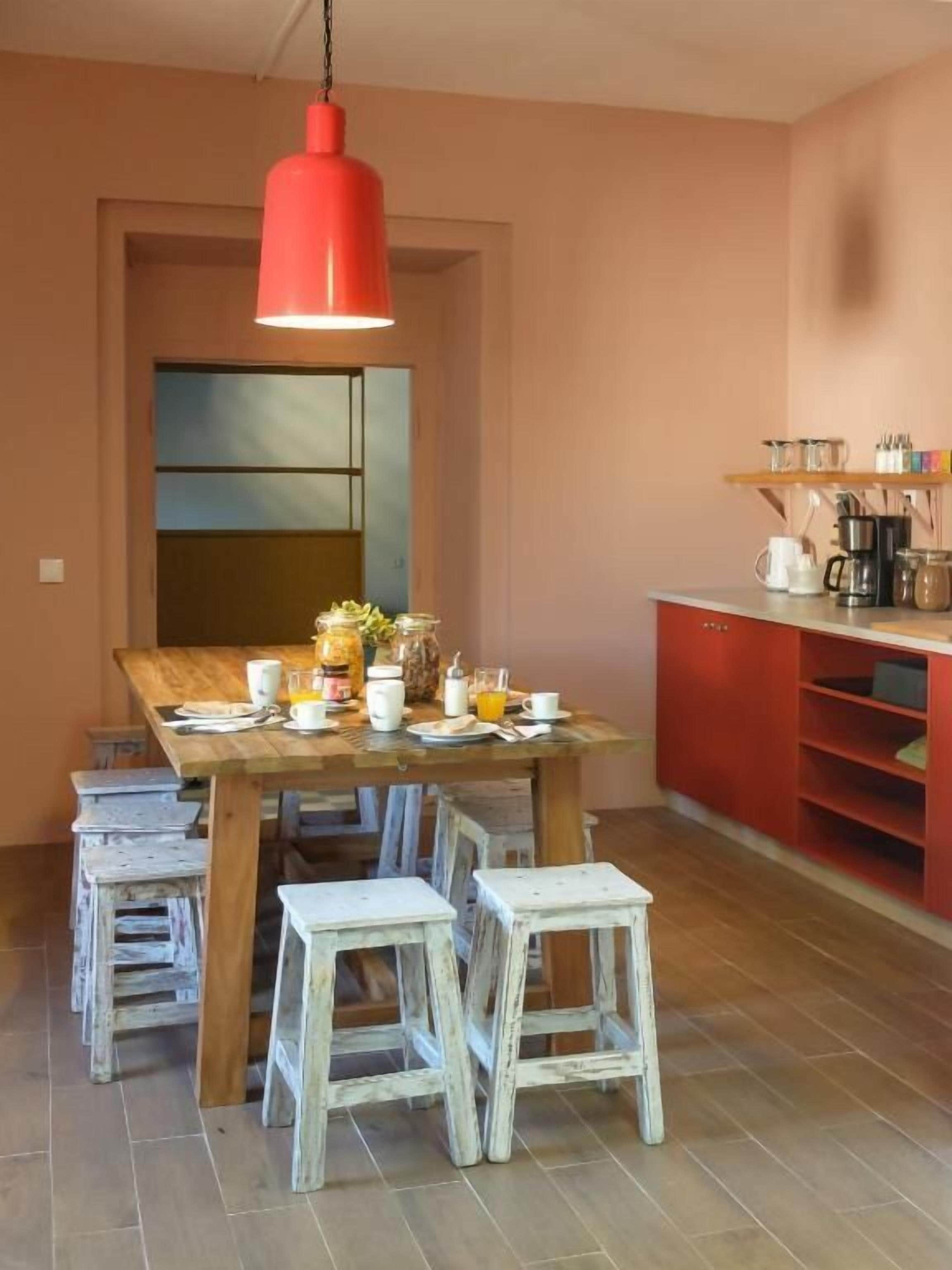 A dining area with a wooden table, white chairs, a red pendant light, and a kitchen area with red cabinets and shelves.