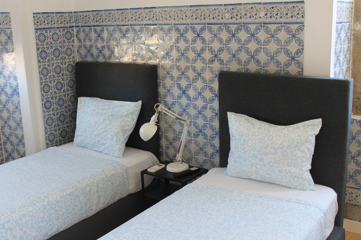 Habitación de hotel con dos camas, una mesita de noche, una lámpara y una pared con diseño azul y blanco.
