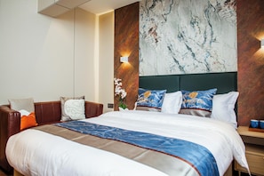 A hotel room with a large bed, a brown leather sofa, a marble wall, and a nightstand with a lamp. - Singala Hotel Ulaanbaatar (Ulaanbaatar)