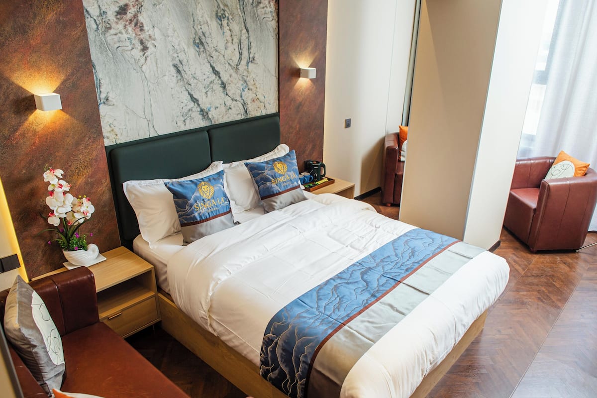 Una habitación de hotel con una cama grande, una mesita de noche, una pequeña mesa de madera, una zona de estar con una silla y una pared de mármol.