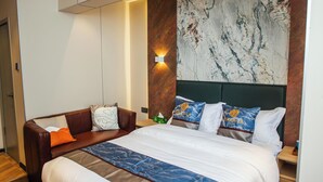 Een hotelkamer met een groot bed, een bruine leren bank, een marmeren wand en een nachtkastje met een lamp.