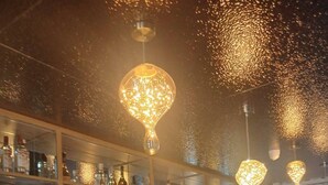 Een bar met planken vol flessen en glazen, een marmeren aanrecht en hangende verlichtingsarmaturen.
