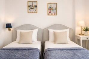 Hotellihuone, jossa on kaksi sänkyä, yöpöytä, lamppu ja kaksi taideteosta, jotka kuvaavat Pariisia ja Lontoota.