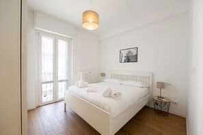 Ein helles Schlafzimmer mit Bett, Nachttisch mit Lampe, Heizkörper und einem Fenster mit Vorhängen.