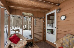 Eine Holzhütte mit einer Glastür, einer Wanduhr und Blick auf einen verschneiten Wald mit Hirschen.