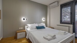 Een netjes opgemaakt bed met witte lakens en een blauw kussen, een nachtkastje met een lamp, en een raam met zonwering.