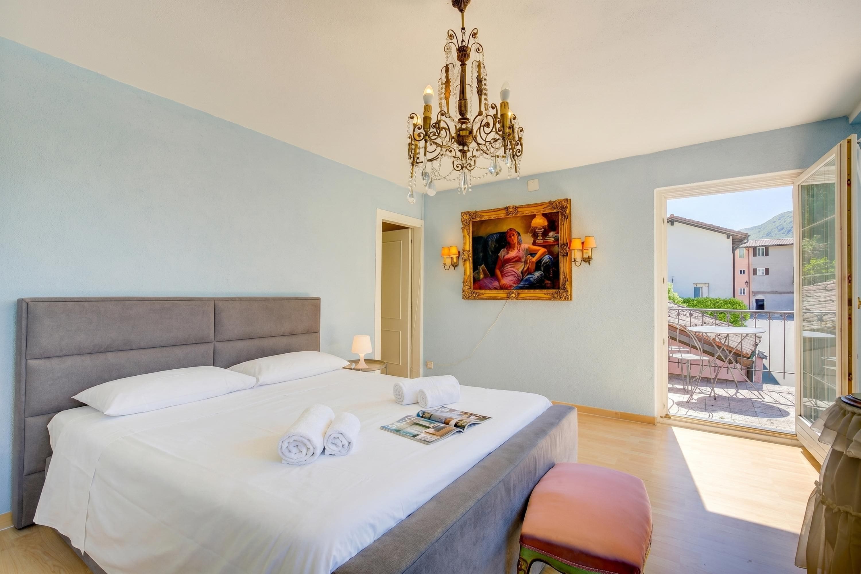 Ein Schlafzimmer mit einem großen Bett, einer Chandelier, einem Gemälde und einem Balkon mit Gartenmöbeln.