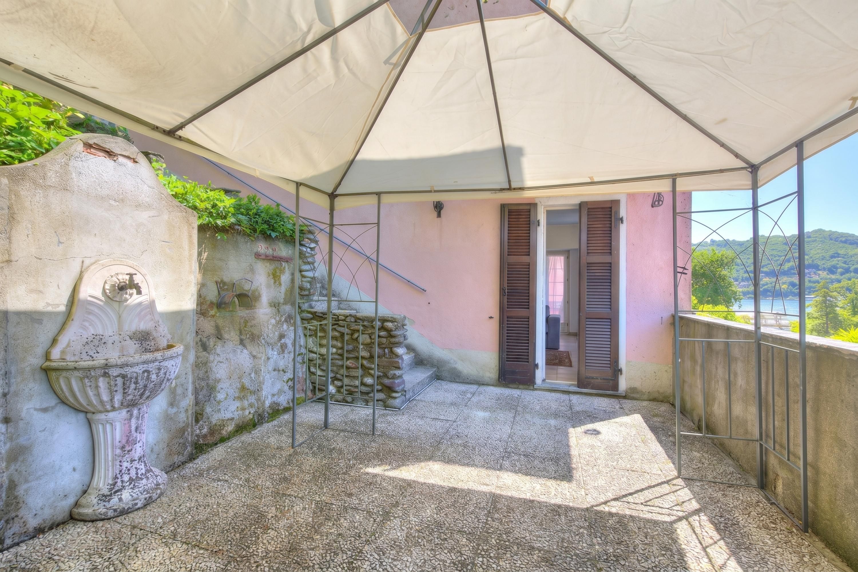 A covered outdoor area with a stone floor, a decorative urn, a metal gate, and a pink wall with a door.
