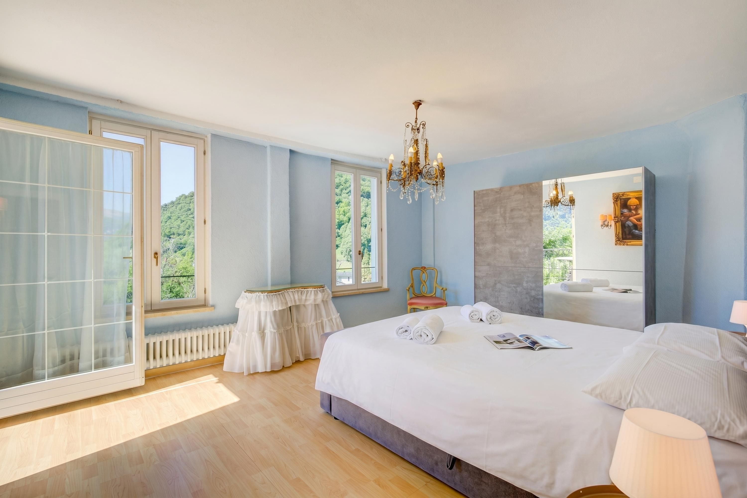 A bedroom with a large bed, a chandelier, a window with a view, a radiator, and a small table.