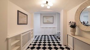 A hallway with a black and white checkered floor, a white radiator, a framed artwork, a ceiling light fixture, and a round mirror. - Brass & Shaker Clapham (London)