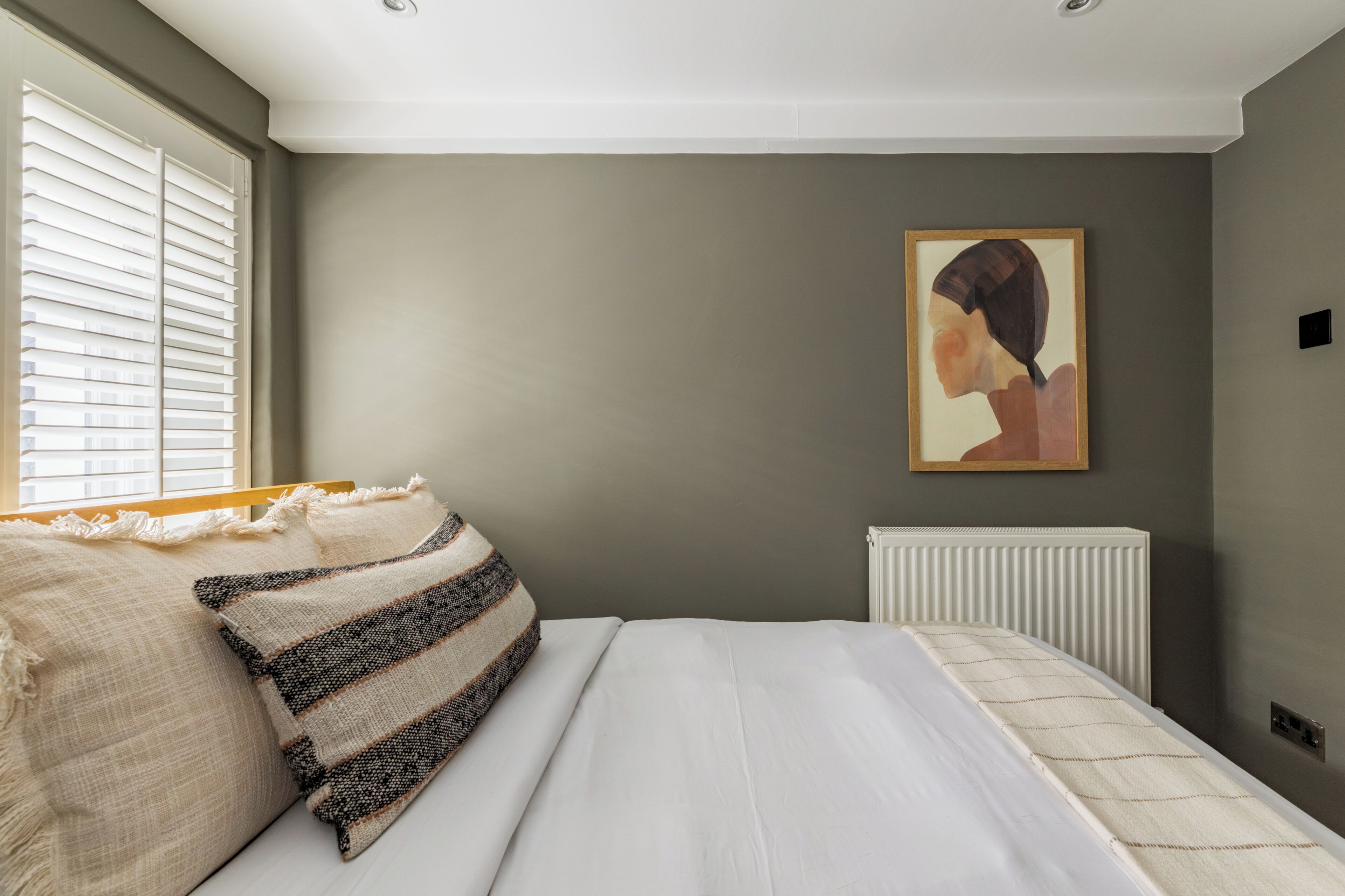 Un dormitorio con cama, almohadas, un radiador y un cuadro colgado en la pared.