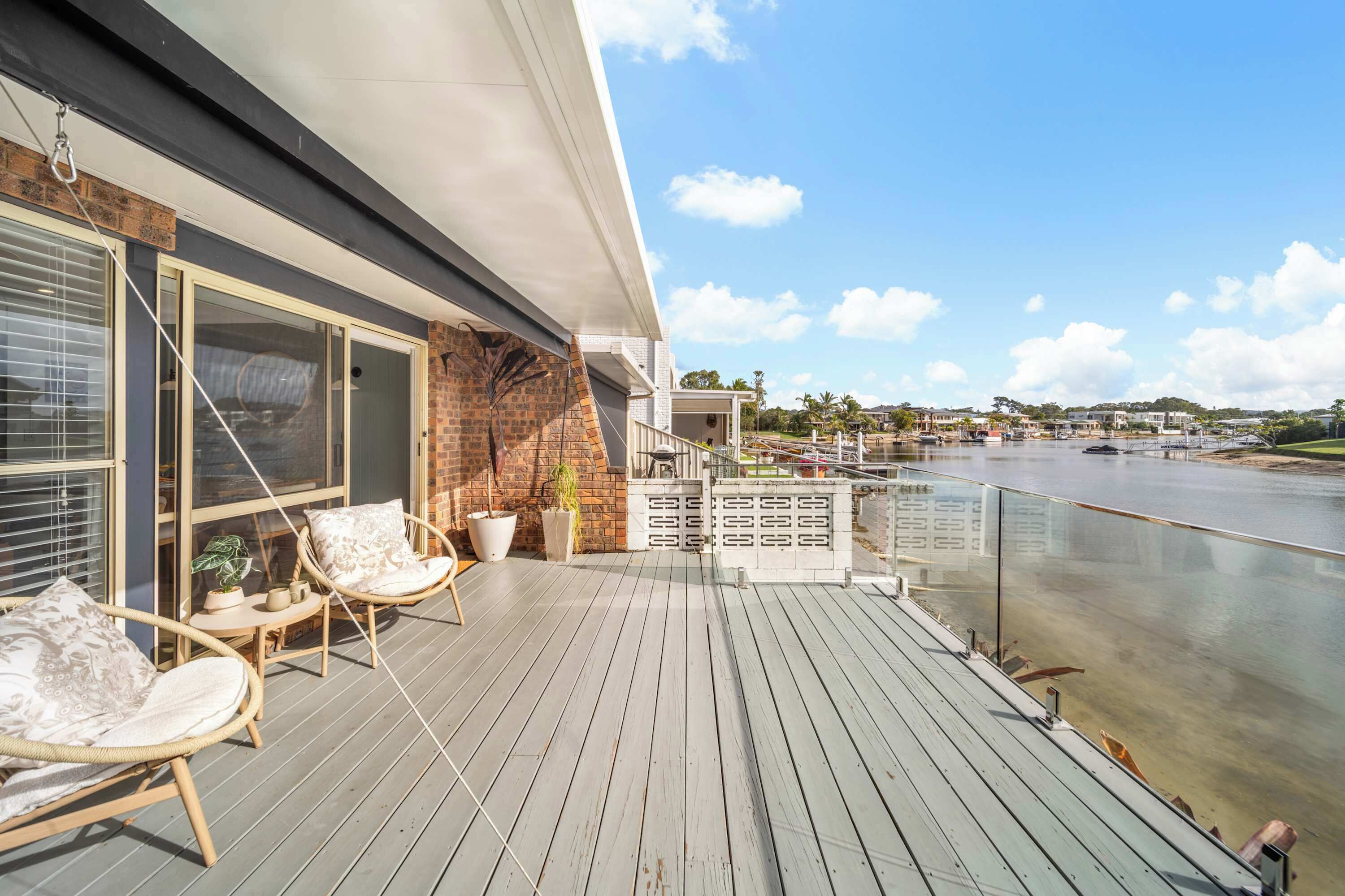 A wooden deck with wicker chairs and a potted plant, overlooking a body of water and buildings.