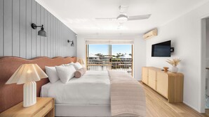 Un dormitorio con cama, mesita de noche, lámpara, cabecera de madera, ventilador de techo, televisor y vistas al exterior.