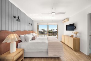Ein Schlafzimmer mit Bett, Nachttisch, Lampe, Holz-Kopfstück, Deckenventilator, Fernseher und Blick ins Freie.