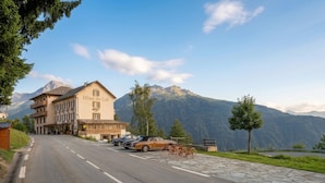 A hotel with a mountain backdrop, a paved road, parked cars, and bicycles. - Hôtel Le Belvédère (Seez)