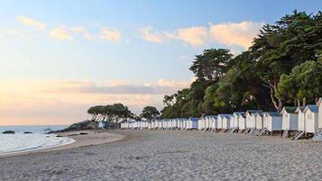 Une plage bordée d’une rangée de cabanes de plage blanches, avec des arbres en arrière-plan et une mer calme.