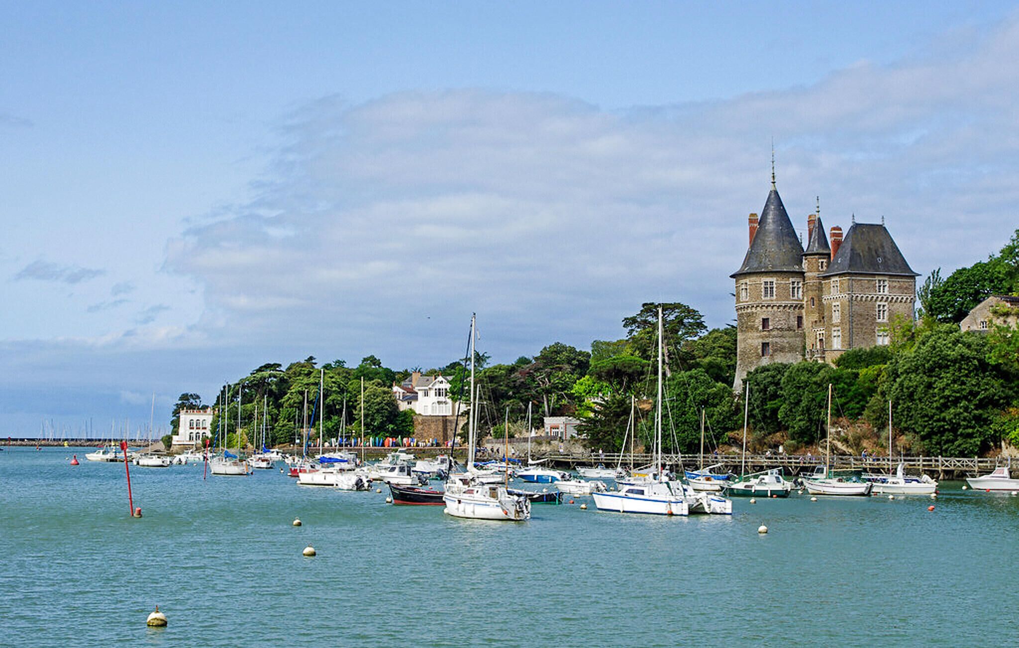 Un paysage côtier avec un château historique, une marina où sont amarrés différents bateaux et une végétation luxuriante.
