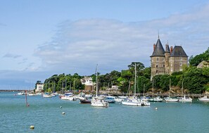 Un paysage côtier avec un château historique, une marina où sont amarrés différents bateaux et une végétation luxuriante.