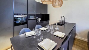 Une cuisine moderne avec des meubles de couleur sombre, un four encastré, un évier et un coin repas aménagé pour quatre personnes, avec une table, des chaises et des verres à vin.
