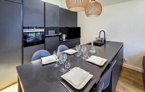 Une cuisine moderne avec des meubles de couleur sombre, un four encastré, un évier et un coin repas aménagé pour quatre personnes, avec une table, des chaises et des verres à vin.