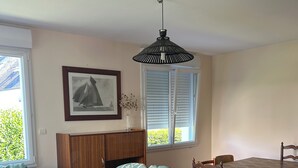 Una habitación con una mesa de comedor de madera, dos sillas de madera, un sofá con tapicería floral, un mueble de madera, un cuadro enmarcado en la pared y una ventana con persianas.