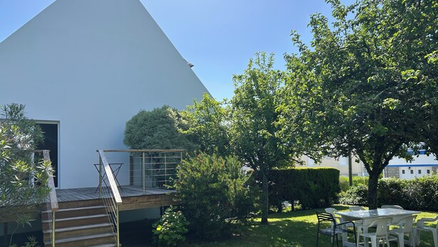 Un edificio moderno con fachada blanca, una escalera de madera, una pequeña zona exterior con mesa y sillas, y un jardín bien cuidado con plantas variadas y un árbol grande.
