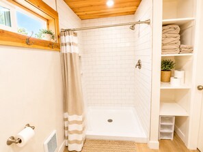 A shower with a glass door, a white tiled wall, a wooden ceiling, and a shelf with towels and toiletries. - Cedar and Stone Studio (San Diego)