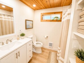 Un bagno con soffitto in legno, lavandino bianco, WC, tenda da doccia e una finestra con vista.