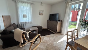 A living room with a sofa, a small table, a TV on a wooden cabinet, and a dining area with chairs.