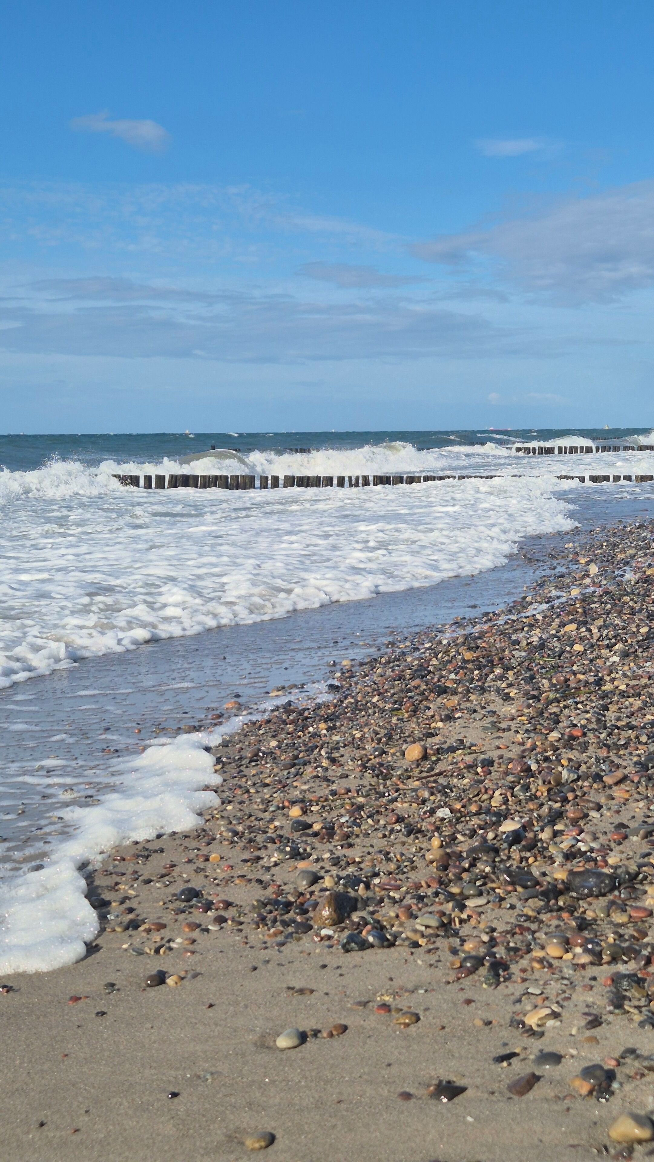 A pebble beach with waves crashing onto the shore and a wooden pier extending into the water.