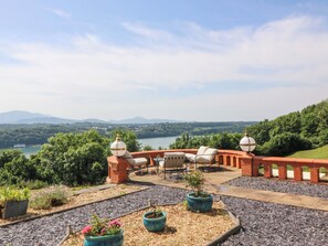 Eine Terrasse mit Gartenmöbeln, Blumentöpfen und einem herrlichen Blick auf einen See und Berge.