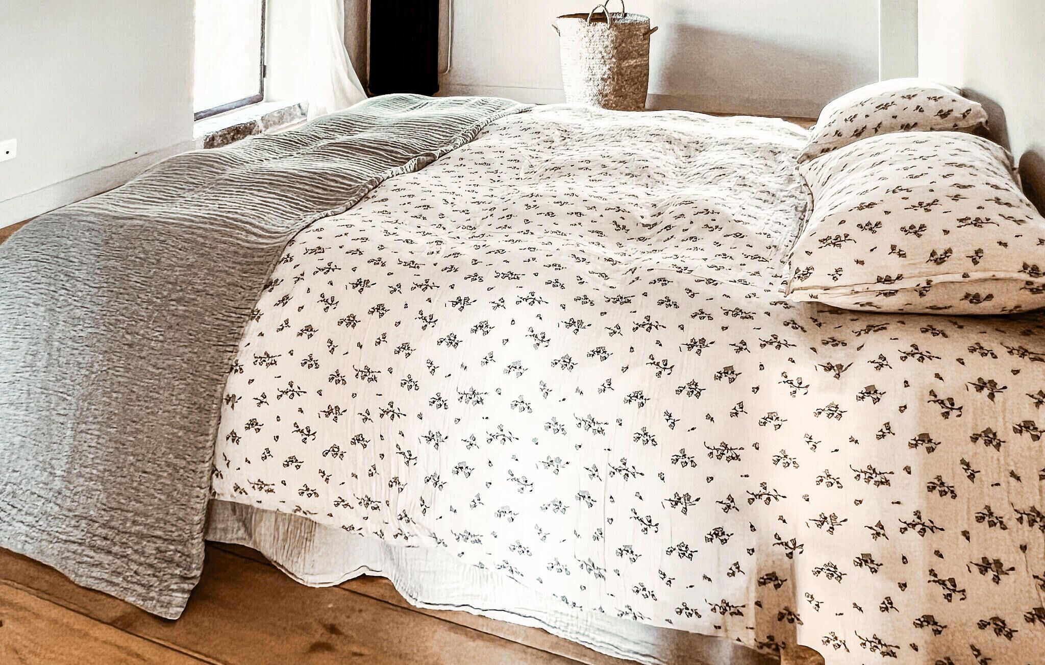 A bed with a floral bedspread, a grey throw blanket, and a woven basket.