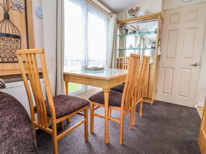 A room with a wooden dining table, four chairs, a glass cabinet with figurines, a radiator, and a window with curtains. - Winchester - 478 (Abergele)