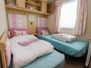 A cozy bedroom with two beds, each with a distinct color theme, a small wooden shelf above, and a window with floral curtains. - Winchester - 478 (Abergele)