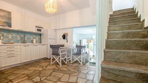 A kitchen with white cabinets, a stone floor, a dining table with chairs, and a staircase. - Possidi Zen Maisonette by RentalsPro (Kassandreia)
