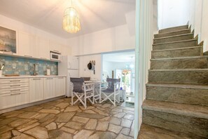 Une cuisine avec des meubles blancs, un sol en pierre, une table à manger avec des chaises et un escalier.