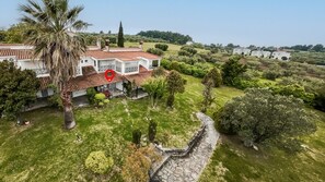 A white villa with a tiled roof, surrounded by lush greenery and a stone wall.