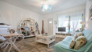 A living room with a stone floor, a round white shelving unit, a television, and a light blue sofa. - Possidi Zen Maisonette by RentalsPro (Kassandreia)