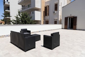 An outdoor seating area with black wicker furniture, a tiled floor, and a white building with balconies and shutters in the background.