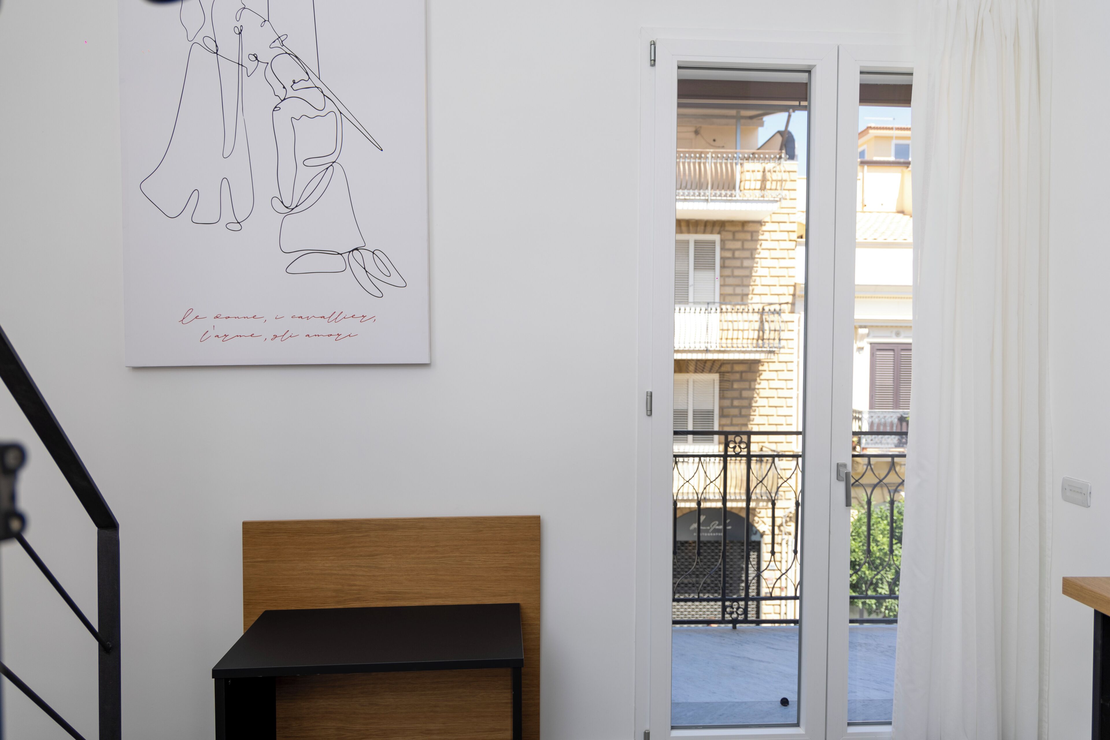 A room with a wooden headboard, a black bench, a white wall with a line drawing, and a door leading to a balcony with a metal railing.
