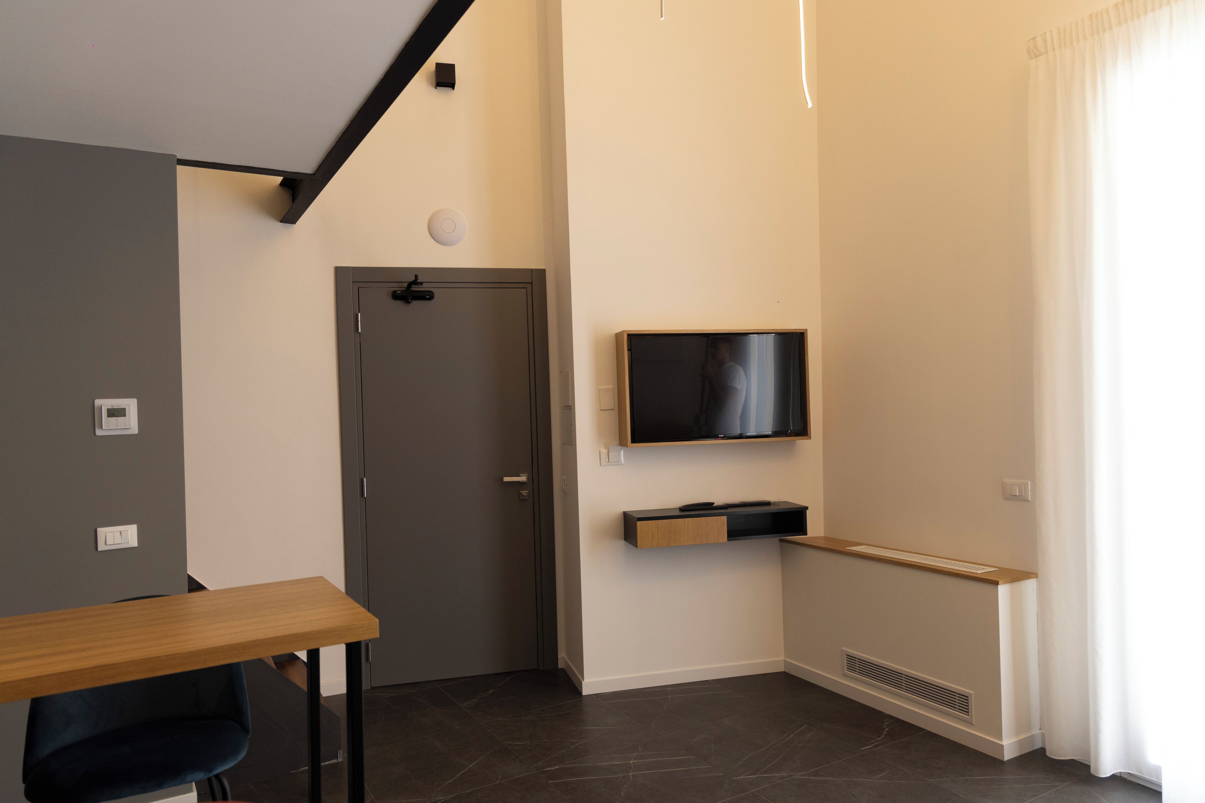 Une chambre moderne avec un bureau en bois, un téléviseur fixé au mur, une porte et une fenêtre avec des rideaux.