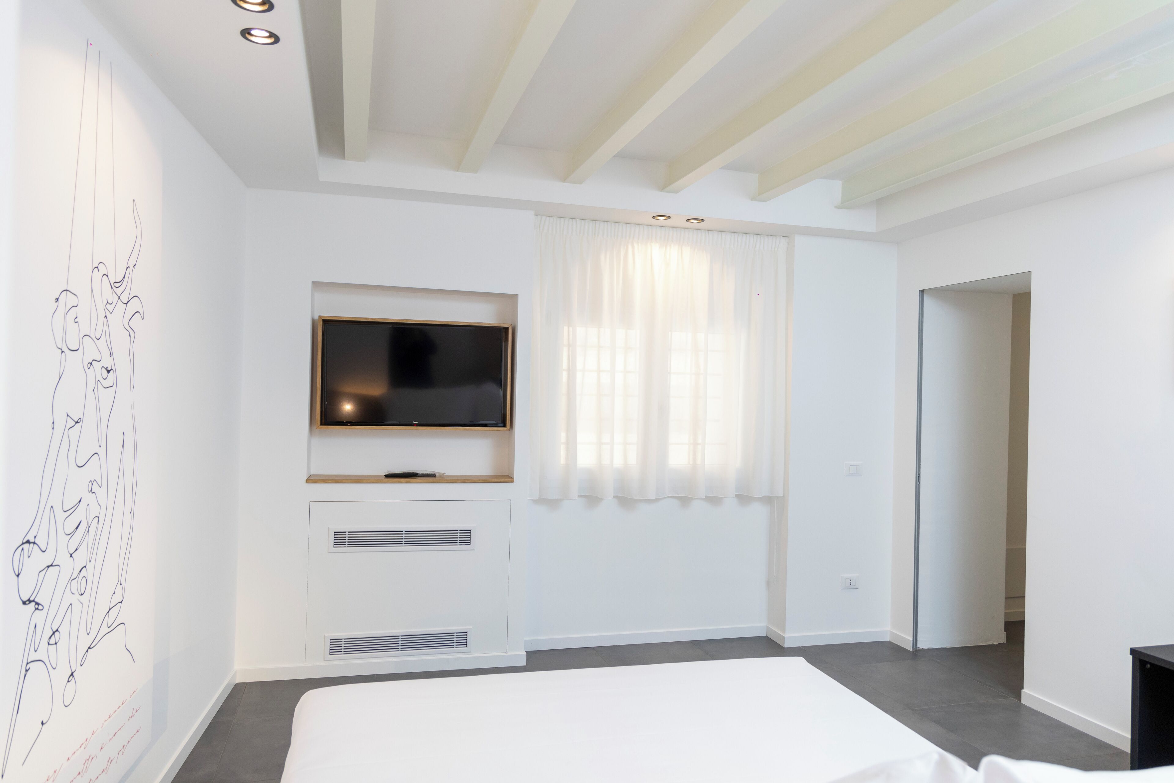 A modern hotel room with a large bed, a flat-screen TV mounted on a white wall, a ceiling with exposed beams, and a window with sheer curtains.