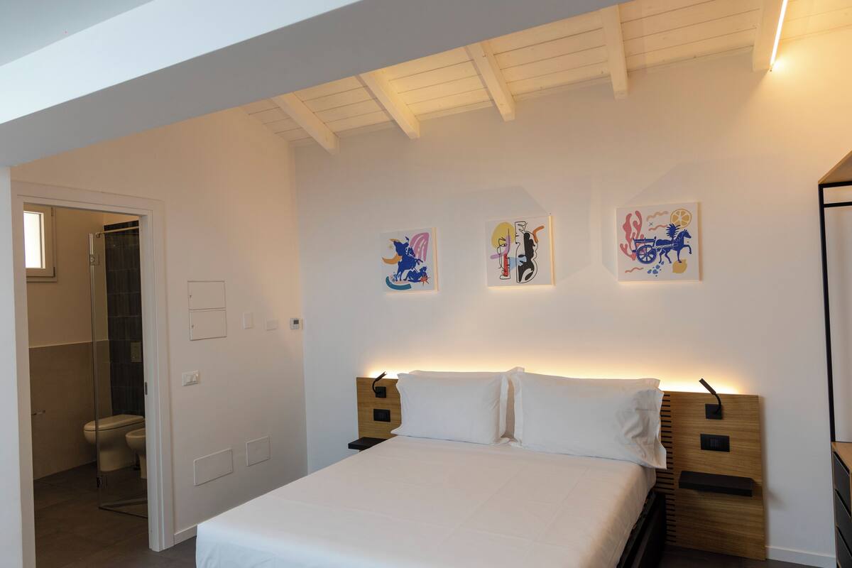 A bedroom with a bed, a nightstand, a wall-mounted lamp, and artwork.